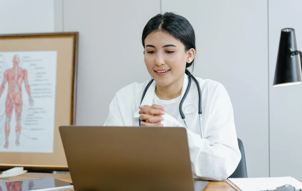 Eine Ärztin sitzt in ihrer Praxis vor einem Laptop und sieht interessiert auf das Display. Die Ärtztin trägt einen weißen Kittel und ein Stethoskop um den Hals.