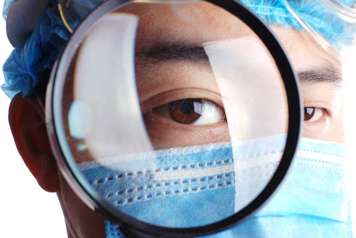 Nahaufnahme einer Person mit chirurgischer Maske und Haube, die durch eine Lupe blickt. Der Fokus liegt auf dem Auge und vermittelt Aufmerksamkeit und Detailgenauigkeit.