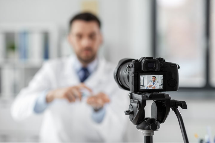 Ein Arzt im weißen Kittel gestikuliert, während er von einer Kamera auf einem Stativ aufgenommen wird – eine professionelle oder lehrreiche Videoaufnahme.